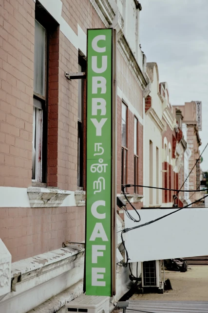 A green sign for a curry cafe is mounted on a red brick building with white trim under a cloudy sky in an urban environment.