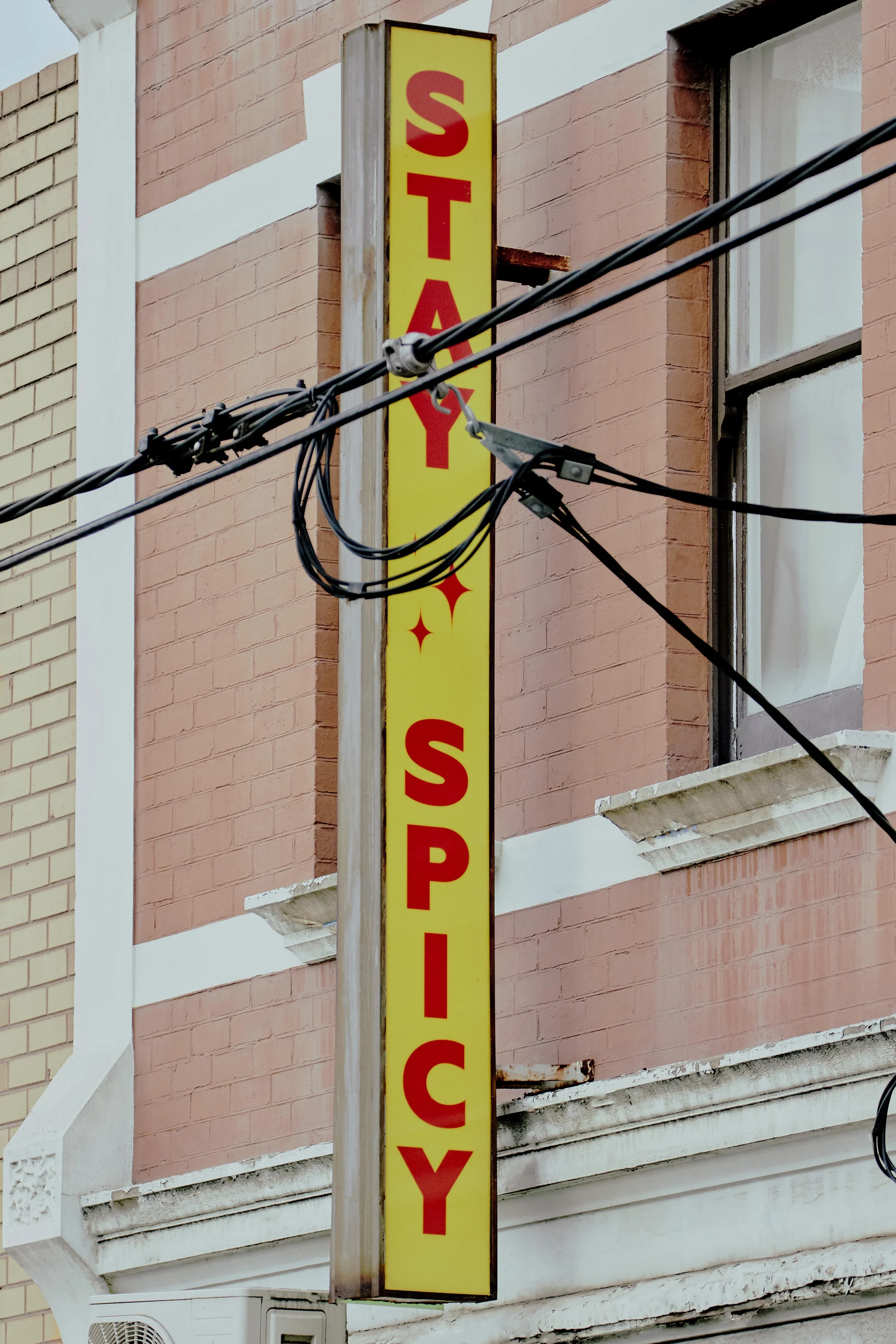 Illuminated vertical sign displaying 'Stay Spicy' in red letters against a yellow background, affixed to a brick building.