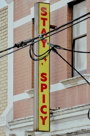 Illuminated vertical sign displaying 'Stay Spicy' in red letters against a yellow background, affixed to a brick building.