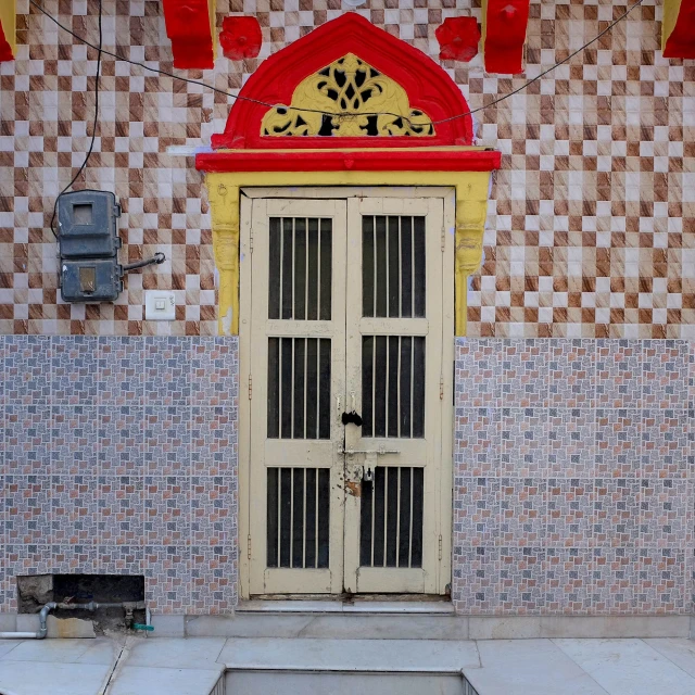 A beige-colored barred door with red and yellow frame stands in front of a patterned tiled wall in an indoor setting.