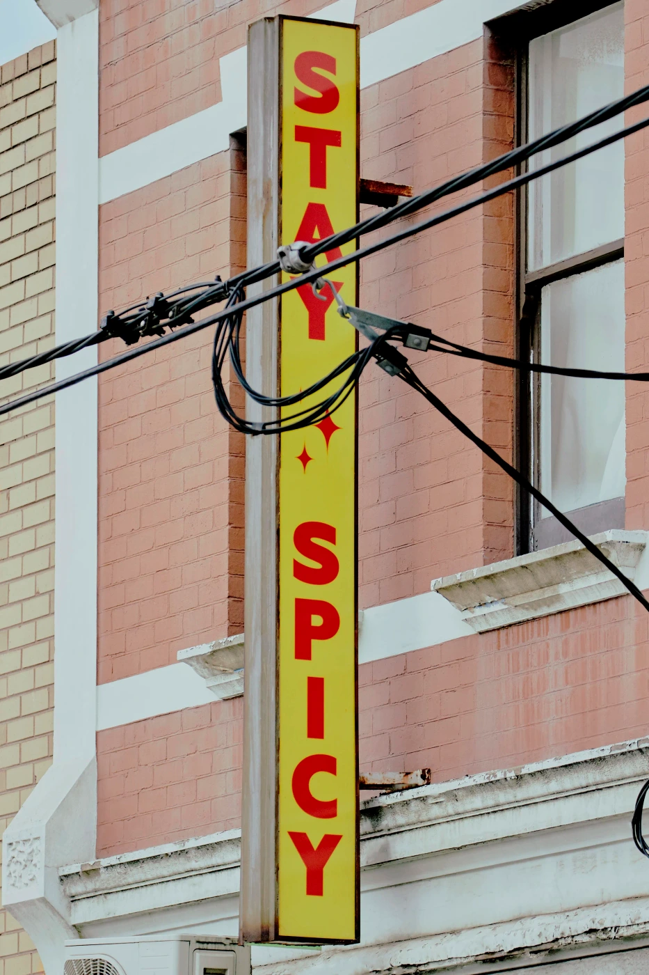 Vertical yellow sign with red vertical text stating Stay Spicy mounted on a pink brick building exterior