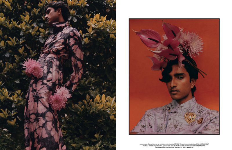 Fashion editorial image featuring a South Asian man in a floral patterned suit with a flower embellishment, posing outdoors among green foliage.