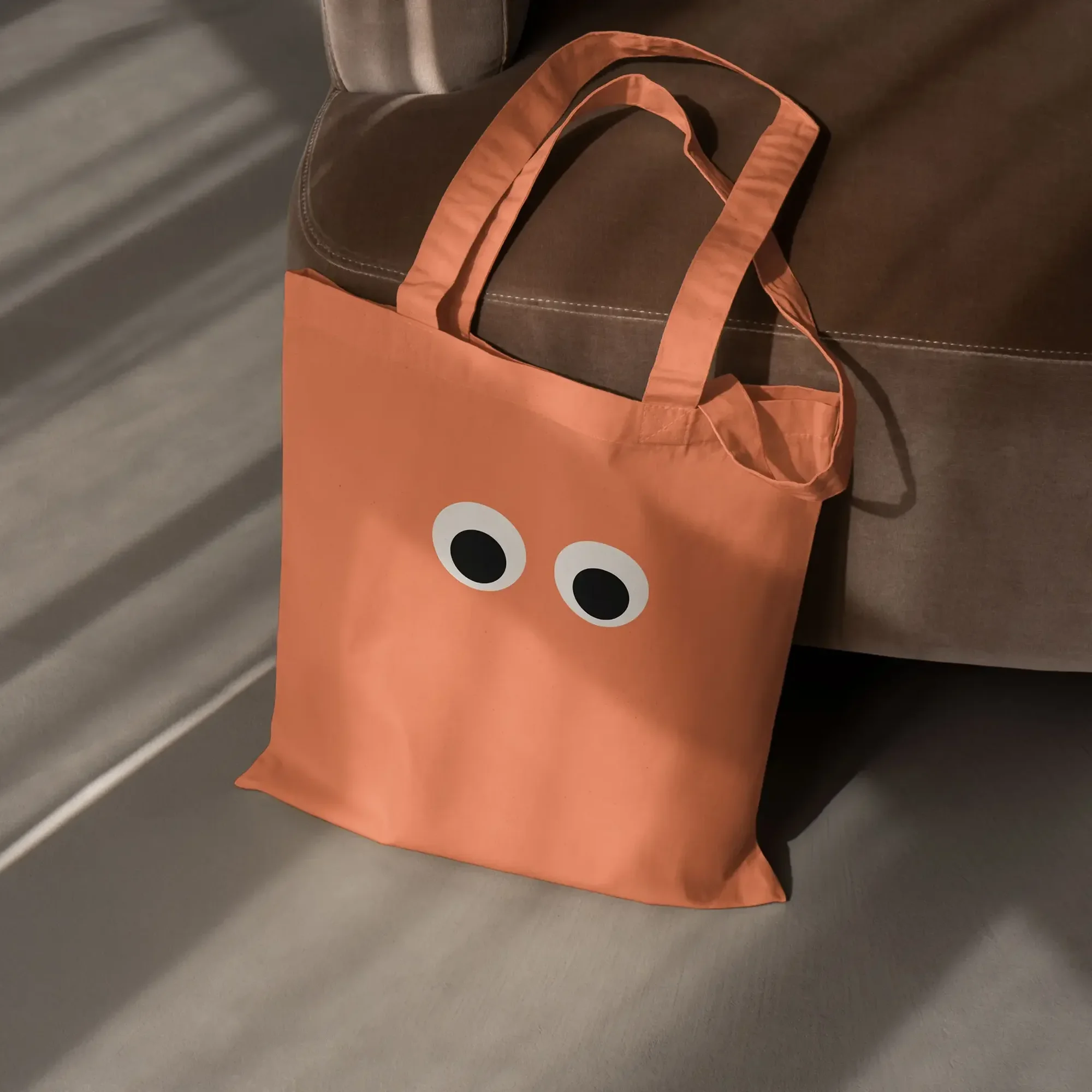 A coral-colored tote bag with cartoon eyes sits on a grey surface next to a brown chair, casting shadows in an indoor setting.