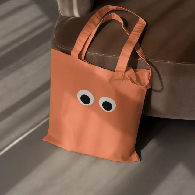 A coral-colored tote bag with cartoon eyes sits on a grey surface next to a brown chair, casting shadows in an indoor setting.