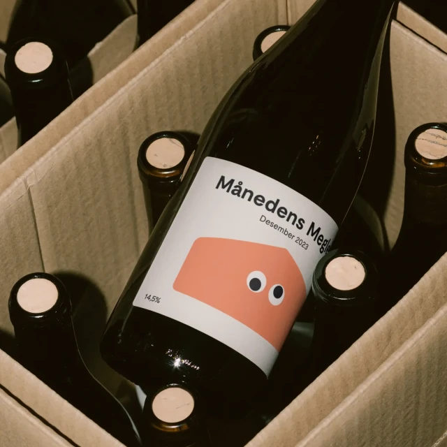 Close-up of a dark wine bottle with a white label featuring an orange graphic with googly eyes, nestled in a cardboard container with other bottles.