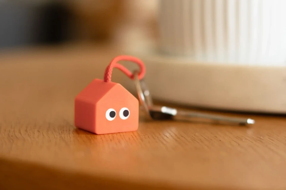 Close-up of an orange house-shaped keychain with googly eyes on a wooden surface, with a key and blurred background element.