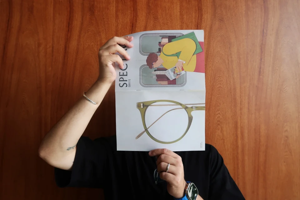 Person holding a magazine cover with eyeglass illustration over their face, against a wooden background, focus on magazine cover.