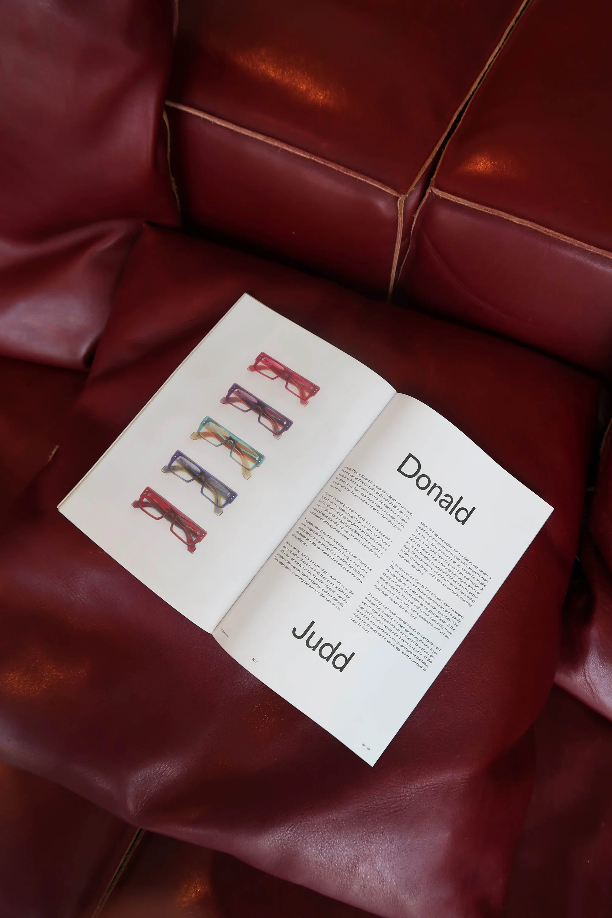 Open magazine featuring eyeglass frames on a red leather sofa showcasing editorial photography and product design in a lifestyle setting.