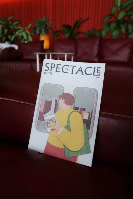 Magazine cover on red leather couch showcases illustration of person on train reading magazine, indoor setting with plants in background.