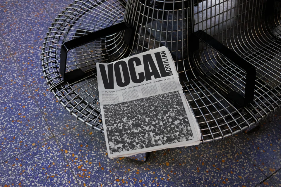 A newspaper titled 'Vocal Civilian' sits on a modern wire chair against a speckled blue and tan floor, capturing a study in contrasts.