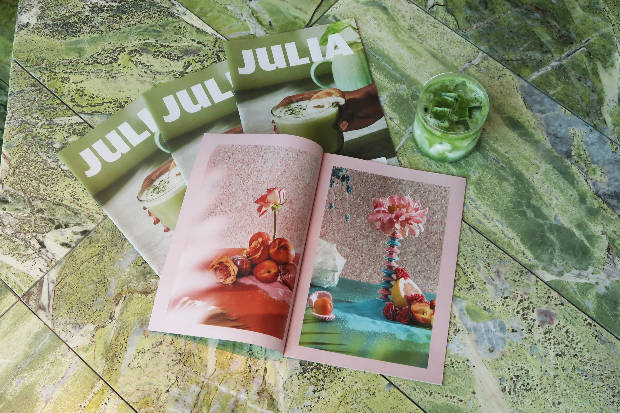 Artistic flat lay of 'Julia' magazines, an open book displaying floral still life, and a matcha drink on a green marble surface.