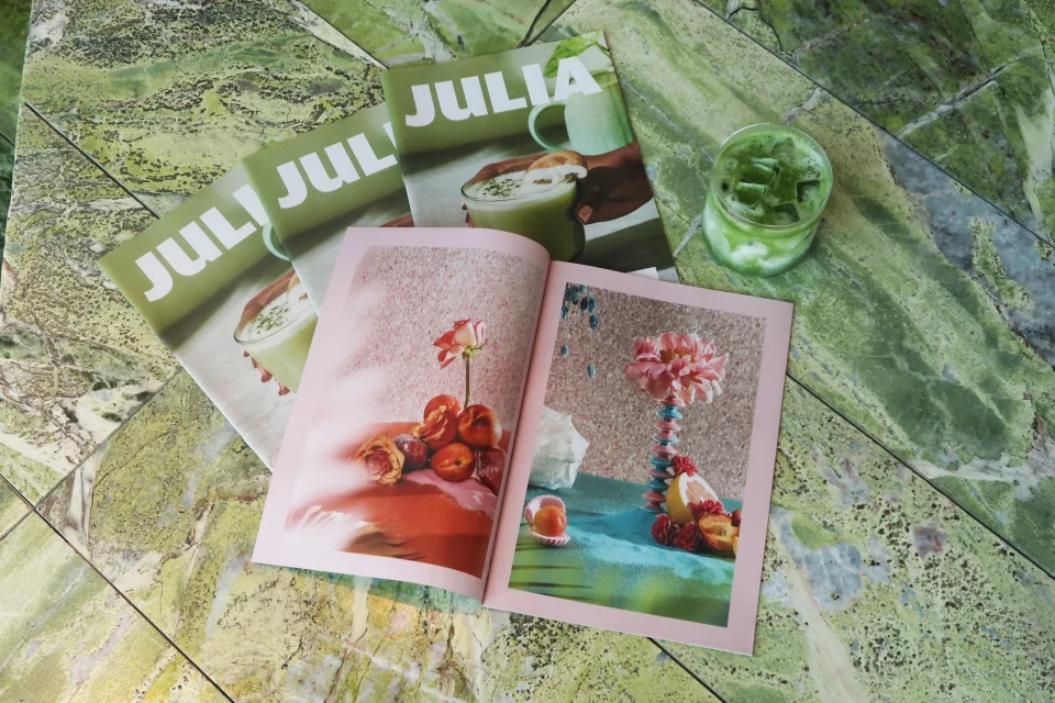Artistic flat lay of 'Julia' magazines, an open book displaying floral still life, and a matcha drink on a green marble surface.