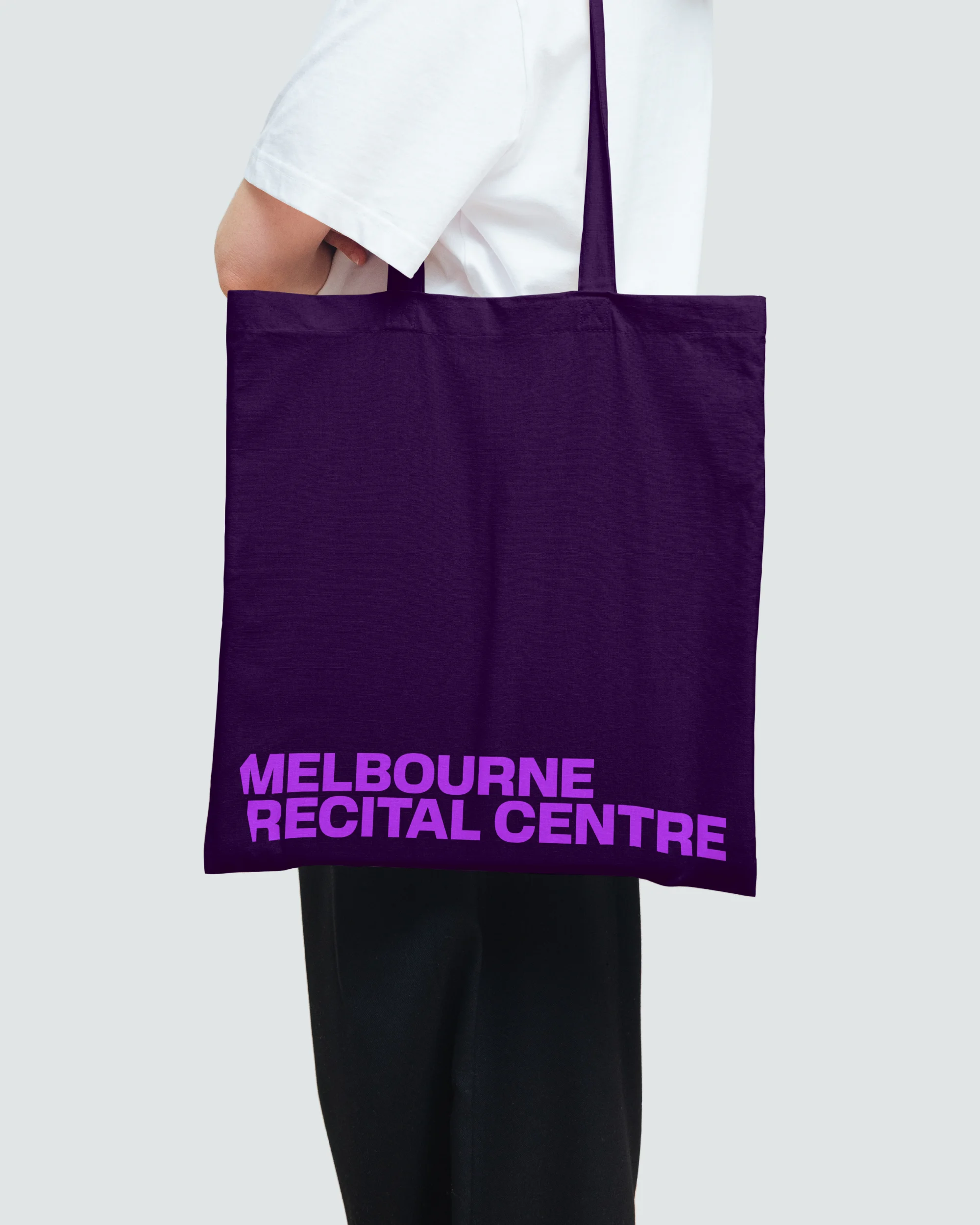 Person carrying a dark purple tote bag with 'Melbourne Recital Centre' printed on it against a light gray backdrop indoors.