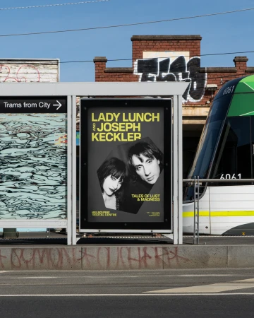 Outdoor shot of a tram stop featuring an advertisement poster of Lady Lunch and Joseph Keckler with a green tram in the background.