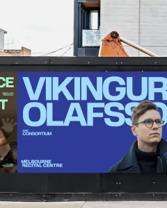Outdoor billboard for Melbourne Recital Centre displaying bold blue typographic poster with portrait of a man wearing glasses