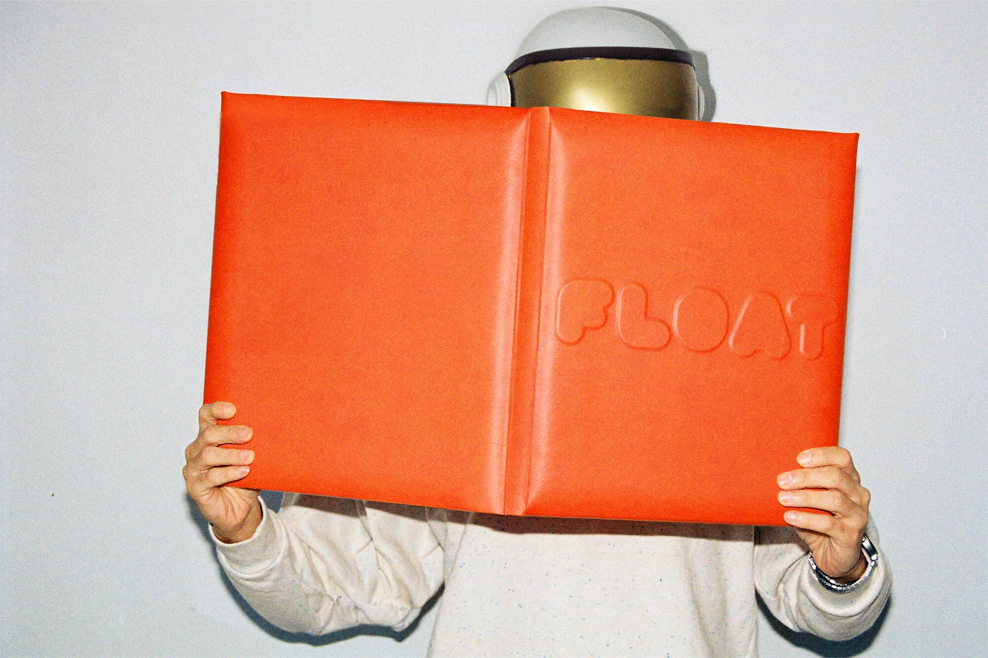 Person in white sweater holding an orange book with embossed letters. A partially visible white helmet is above the book.