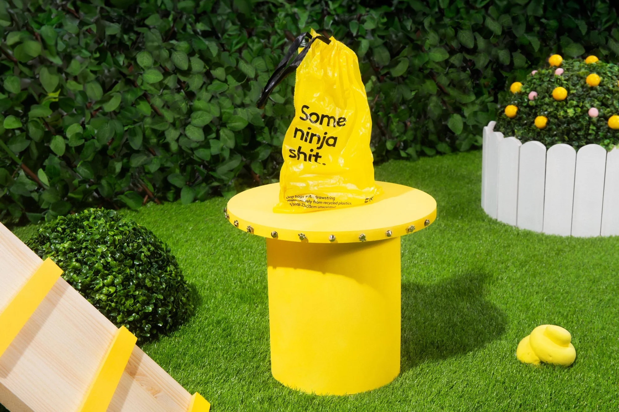 Yellow dog waste bag on a matching yellow dispenser in a green backyard setting with a small mound of fake dog poop.