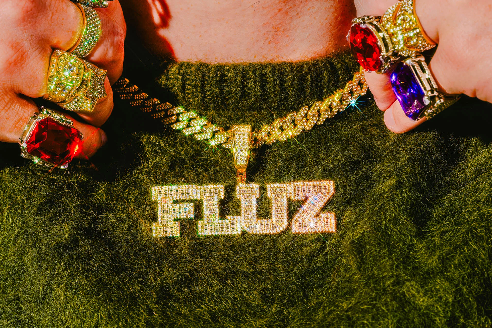 Person wearing a gold chain with a diamond pendant that reads "FLUZ" and multiple large gold rings with colorful gems. 