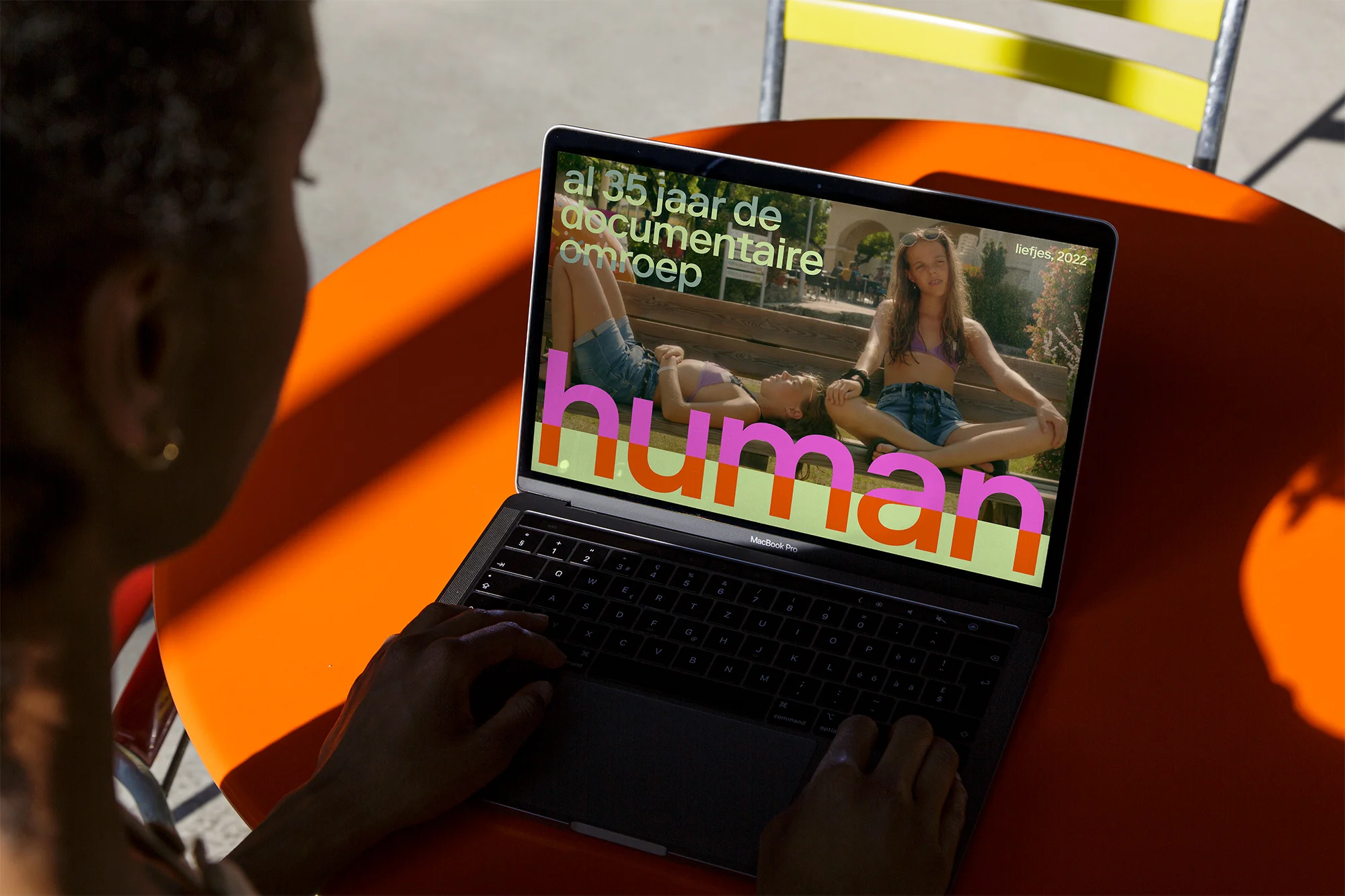 Woman using a laptop to view a documentary featuring two young women on screen; vibrant orange table and yellow chair are visible.