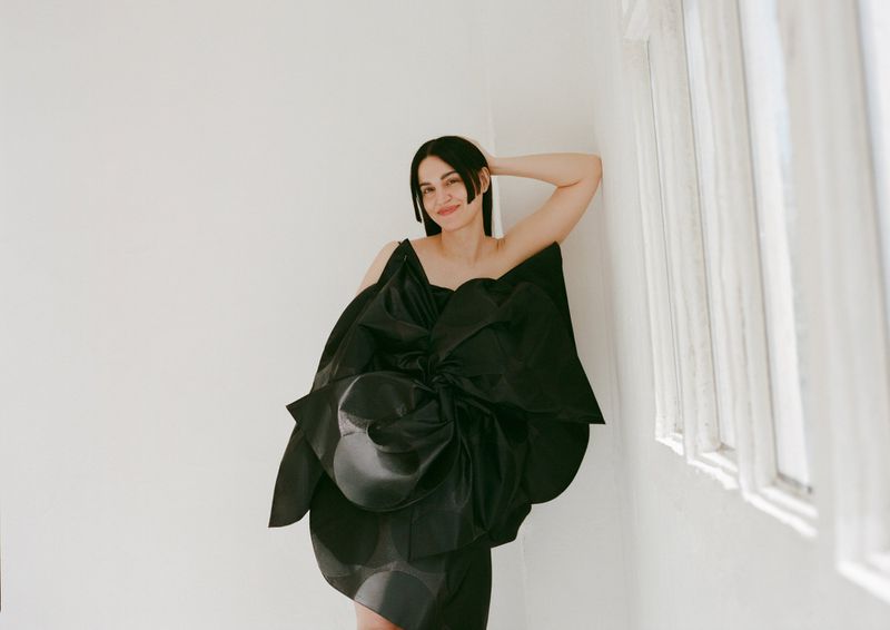 Woman in a dramatic black couture dress with a large bow detail posing indoors against a white wall near a window with natural light.