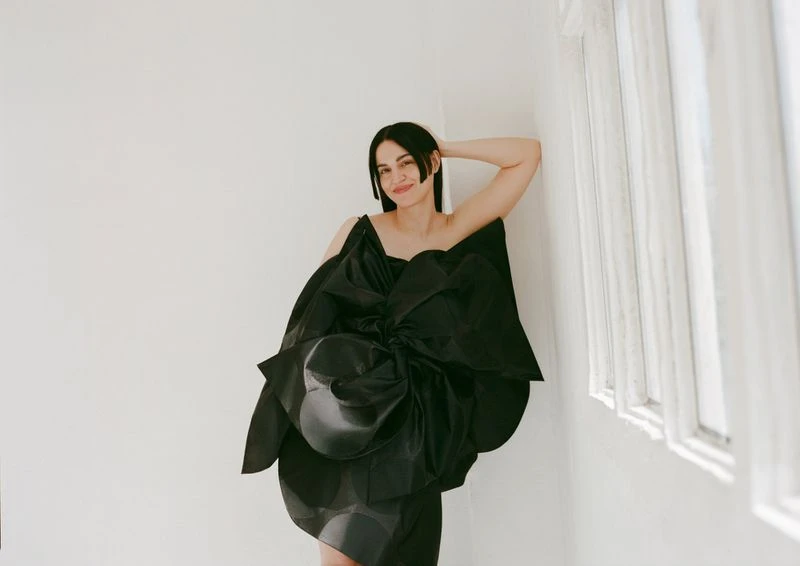 Woman in a dramatic black couture dress with a large bow detail posing indoors against a white wall near a window with natural light.