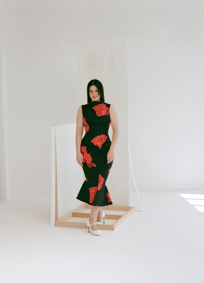 A woman stands in a black, sleeveless, midi dress with a red rose pattern. She wears white heels and poses in a minimal white setting.