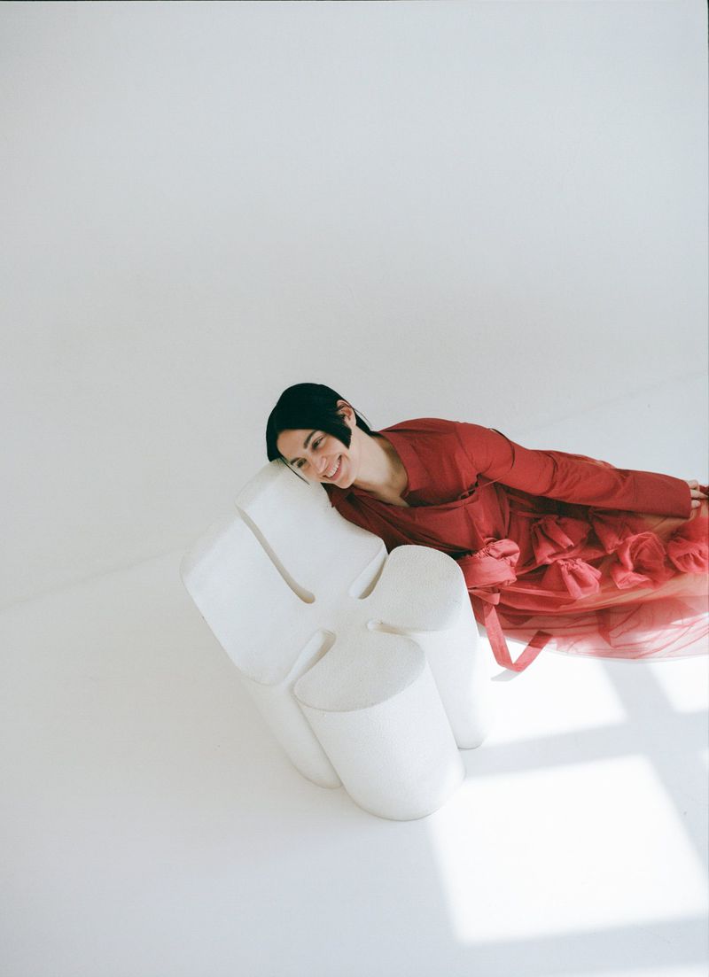 A woman in a red dress poses in a white photo studio, leaning against a sculptural white chair with a bright smile on her face.
