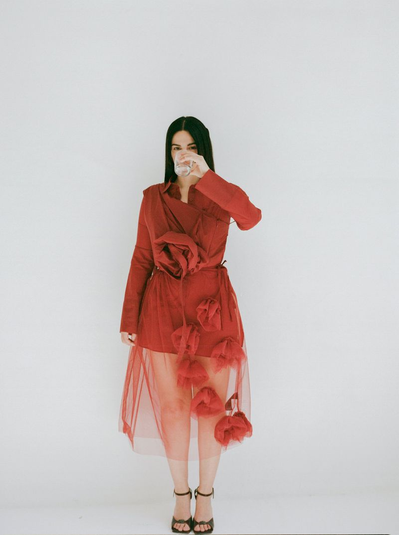A full shot of a woman with long dark hair drinking from a glass. She is wearing a red dress with floral embellishments, against a white backdrop.