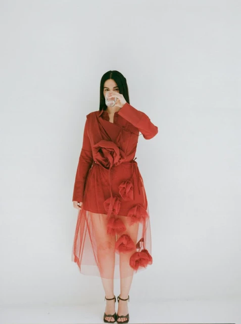 A full shot of a woman with long dark hair drinking from a glass. She is wearing a red dress with floral embellishments, against a white backdrop.