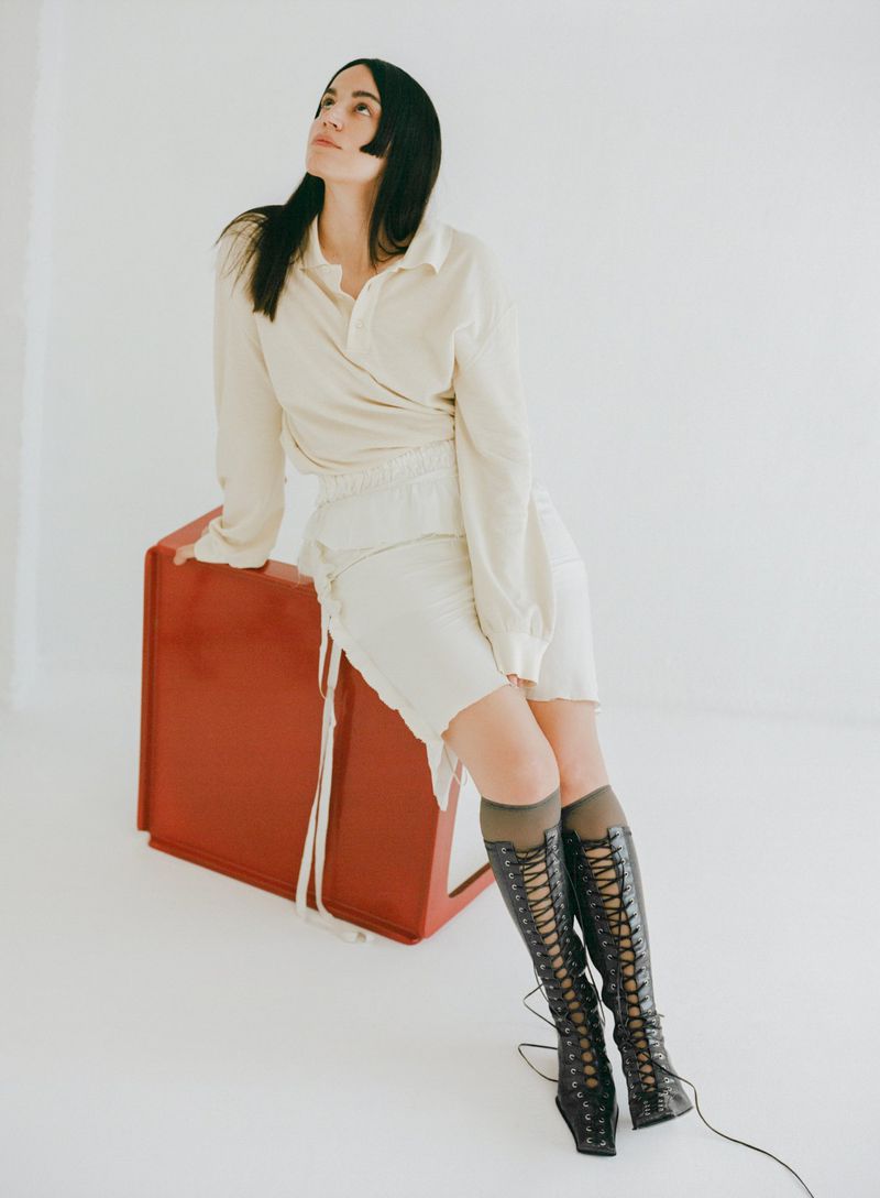 Adult woman in a light-colored wrap shirt and skirt posing indoors against a white background, seated on a red box. She wears high boots.