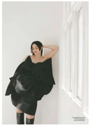 Woman in a black structural dress and tall boots leaning against a white wall next to a window for Vogue Espana