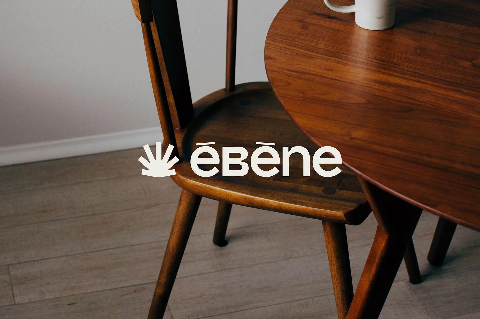 Close up of a wooden chair and round wood table with a coffee mug on top, next to a white wall and light wood flooring.