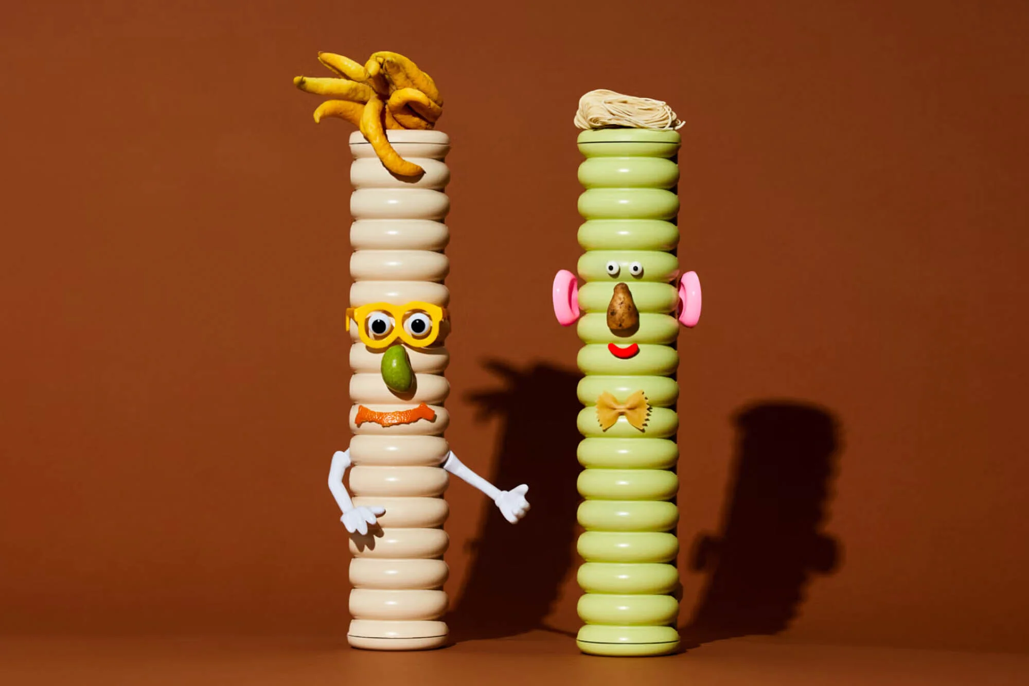Two stacked, colorful columnar characters assembled from fruits, vegetables, pasta, and found objects against a brown background.