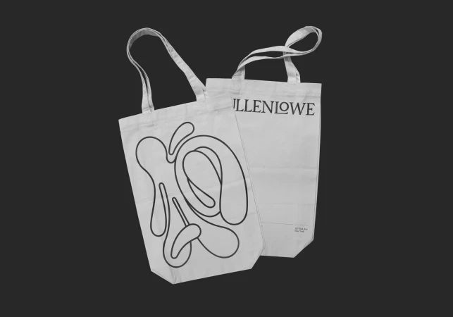Two white tote bags with graphic designs on a dark gray background, one featuring an abstract drawing and the other brand text.