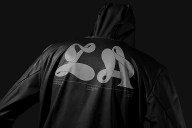 Rear view of a person wearing a black hooded rain jacket featuring large white typography printed across the back against a dark background.