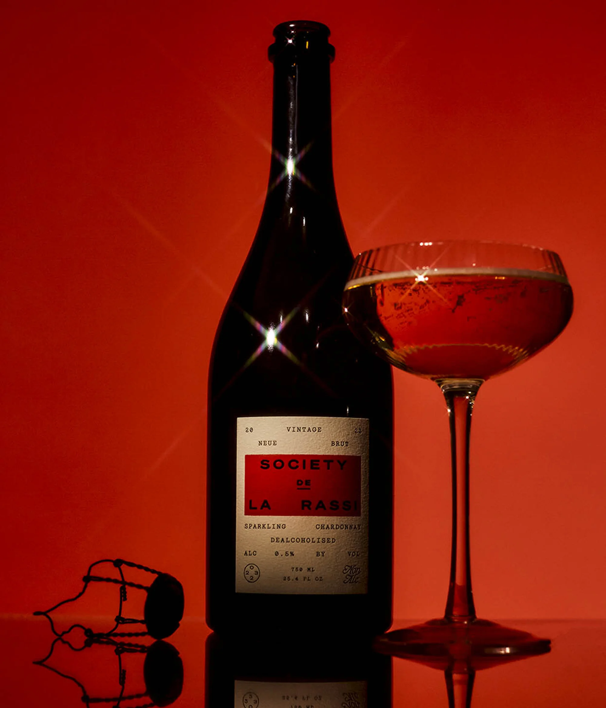 Studio shot of a wine bottle and glass of sparkling rose wine set against a red background with the wine cork lying beside it.