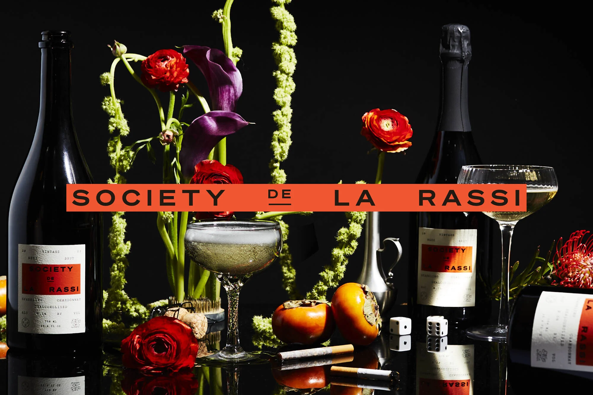 Elegant still life featuring two wine bottles, flowers, fruit, dice, and cigarettes on a reflective surface against a dark backdrop.
