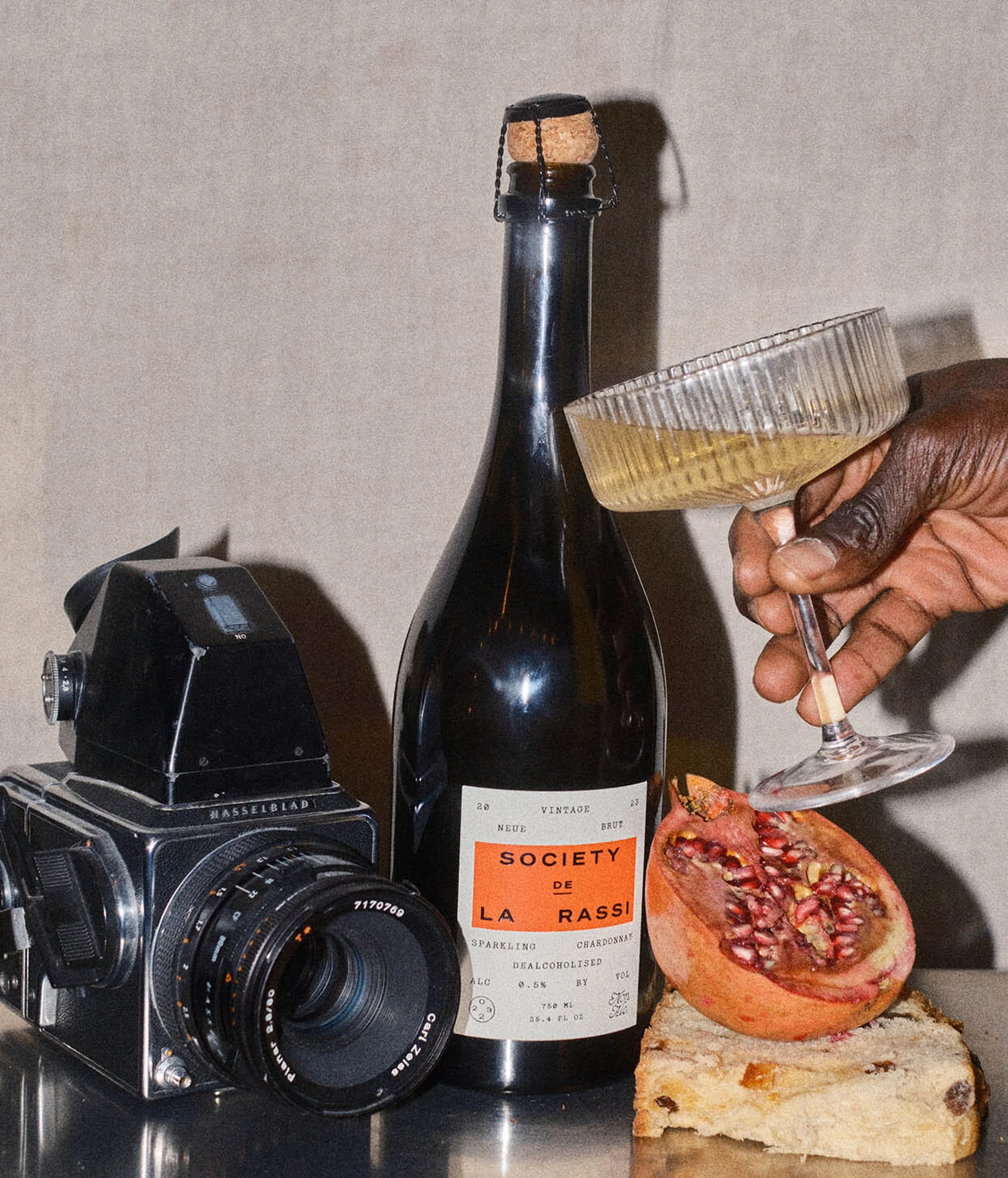 Still life photography features a wine bottle, a wine glass held by a hand, a camera, and a pomegranate against a neutral background.