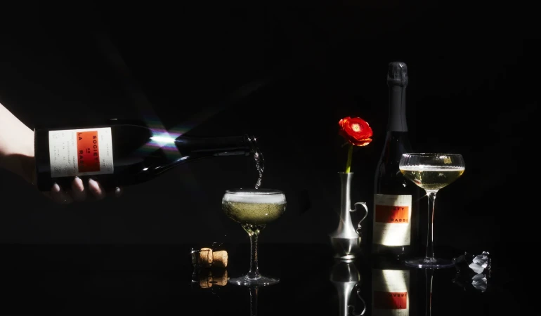 Pouring sparkling wine into a coupe glass on a black reflective surface next to a wine bottle and flower in a dark studio.
