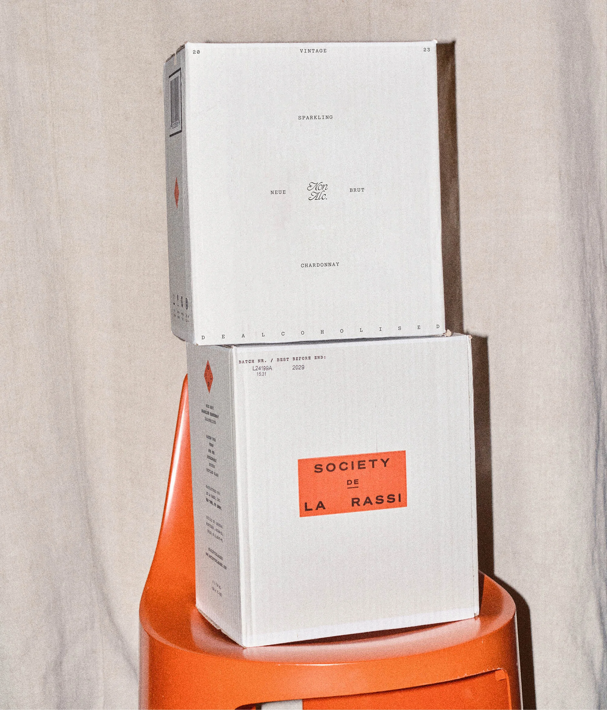 Two stacked white boxes with brand labels sit on an orange stool against a cream backdrop, showcasing minimal product packaging.