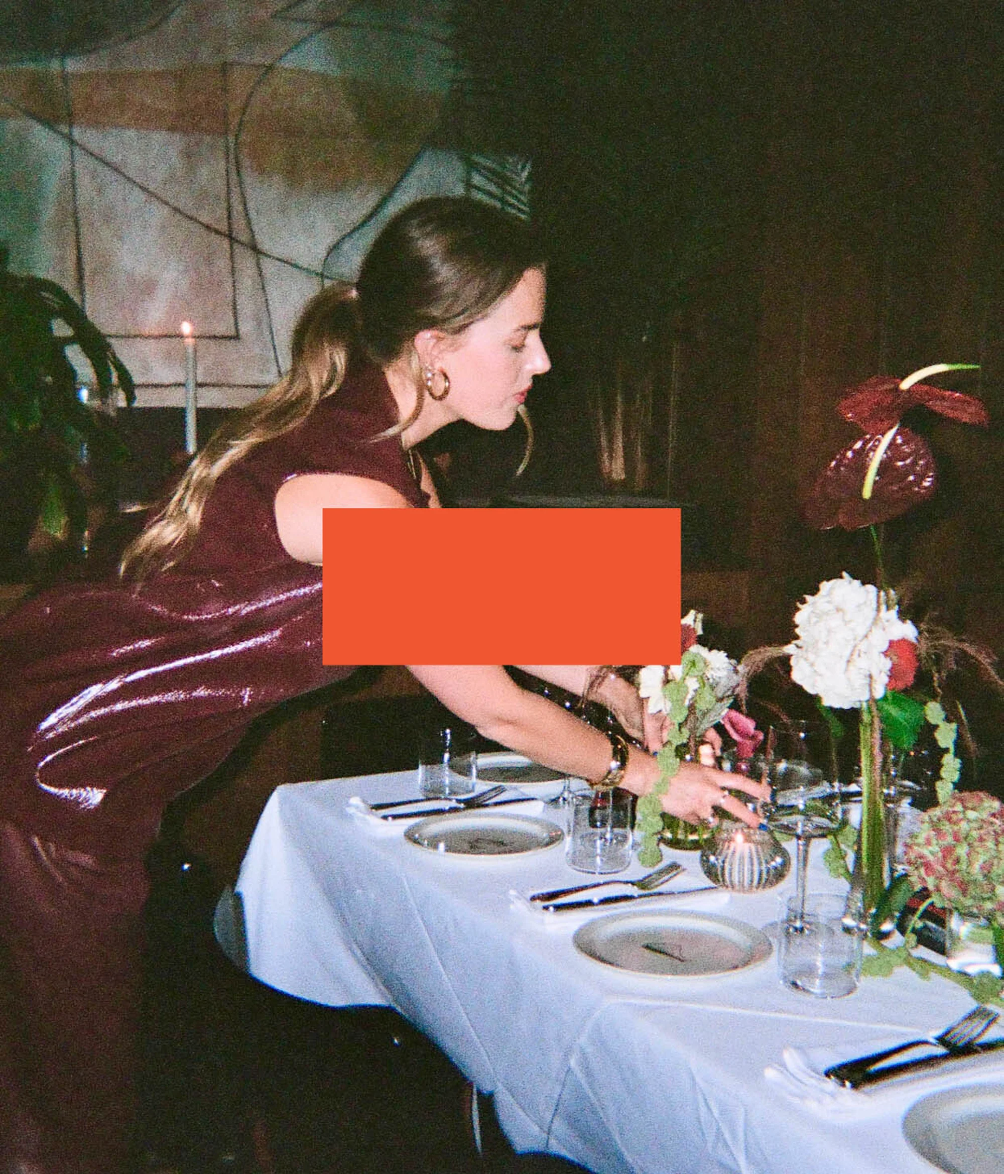 Adult female arranging flowers on a table set with plates and glasses indoors, wearing a maroon sequin dress and gold earrings.