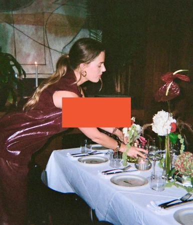 Adult female arranging flowers on a table set with plates and glasses indoors, wearing a maroon sequin dress and gold earrings.