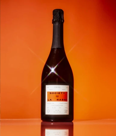 Elegant bottle of Society de la Rasse sparkling wine on a reflective surface, set against a vibrant orange background, evokes luxury.