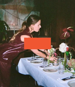Woman in burgundy sleeveless top leaning over a white-set dining table arranged with flower centerpieces and candles.