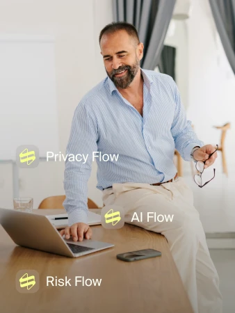 Bearded man in a blue striped shirt leans on a desk, using a laptop in a bright indoor space with natural lighting.