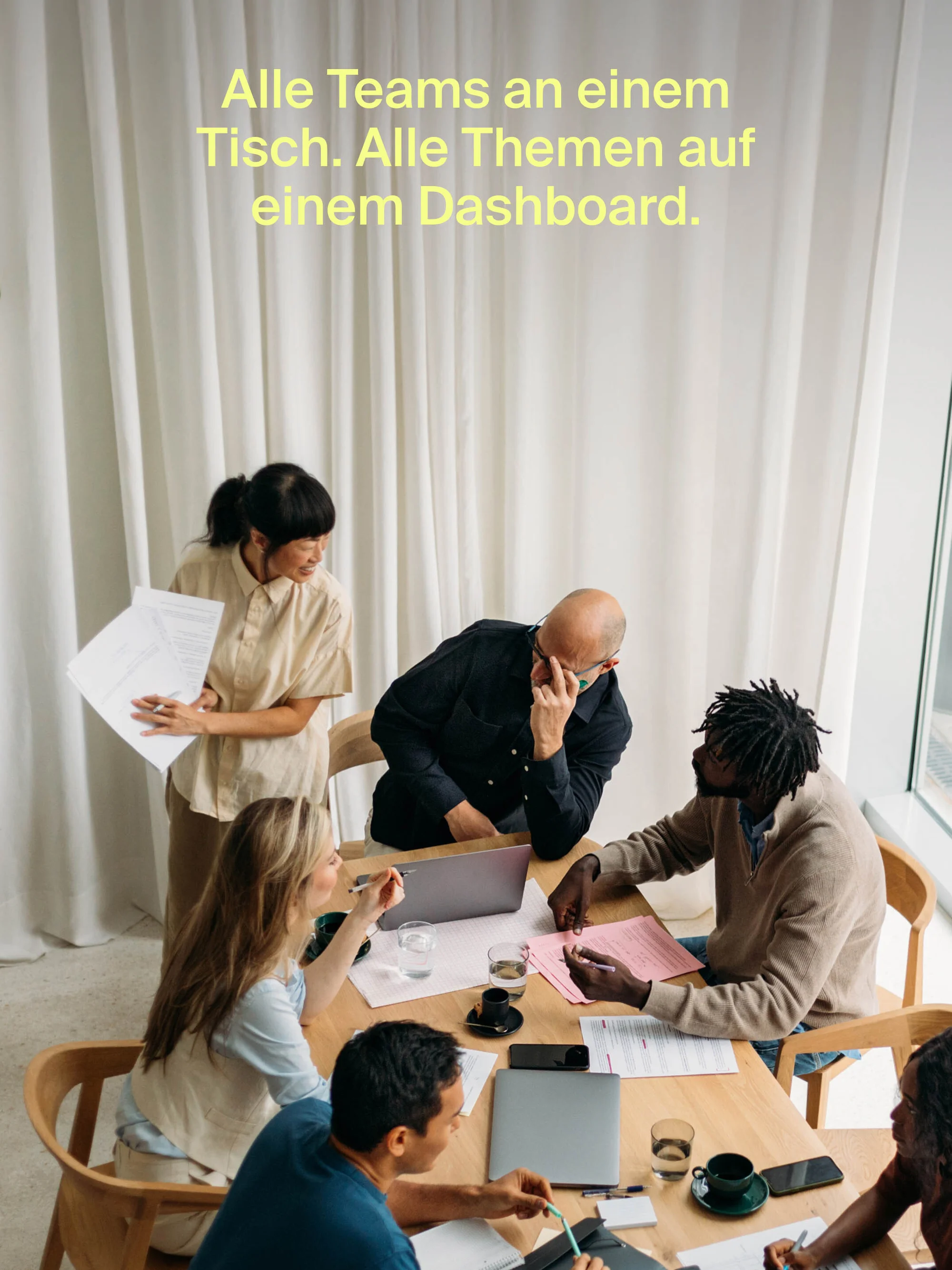 Group of diverse professionals collaborate around a table with laptops and documents in a well-lit office with natural light.