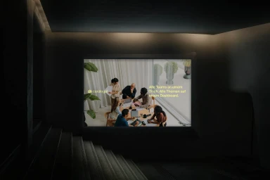 Illuminated digital display screen mounted on a dark architectural wall above a staircase showing a team meeting