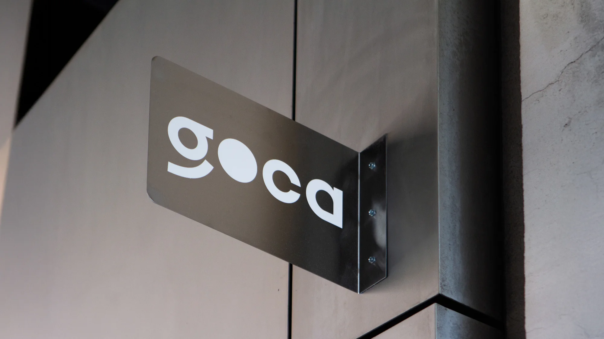 GOCA logo on a metallic sign, mounted on a grey wall with stainless steel brackets and screws, studio environment, close-up shot.