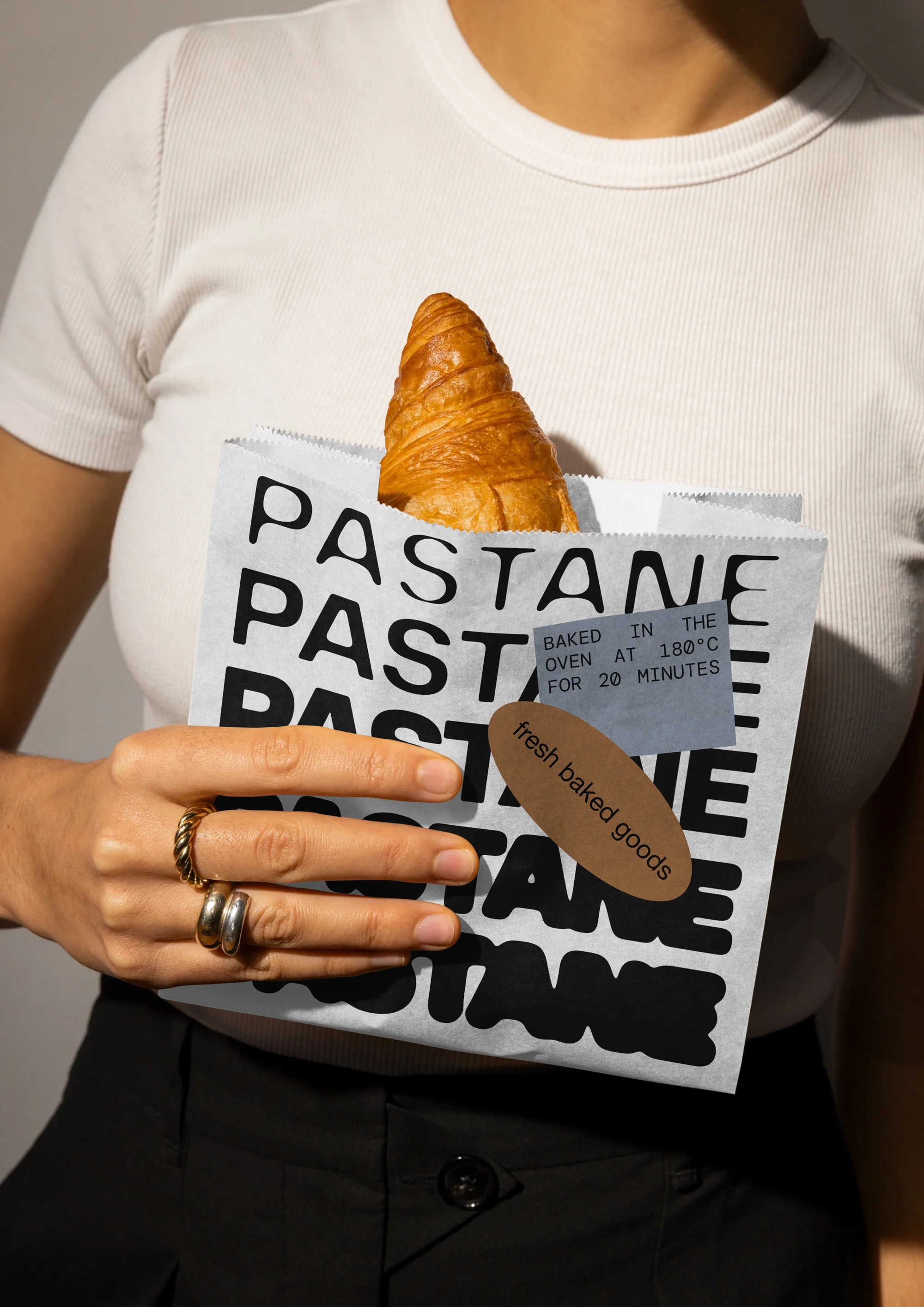 Adult female holds paper bag with fresh croissant, against neutral background. Bag with text ‘PASTANE’, labels ‘baked goods’.