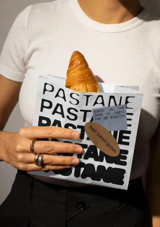 Adult female holds paper bag with fresh croissant, against neutral background. Bag with text ‘PASTANE’, labels ‘baked goods’.